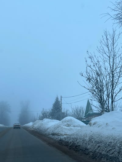 The image displays a winter road scene in Yasenevo, Russia, under foggy or hazy conditions. A dark paved road, appearing wet or soiled, extends into the distance from the lower-center of the frame. A light-colored sedan is observed moving away from the viewer, centered on the left half of the road. Along the right edge of the road, prominent, irregular snowbanks are present, exhibiting patches of dirty brown slush near the pavement. Further right, a large bare deciduous tree stands, partially covered in snow. Behind the snowbanks and trees, a structure with a steep, pointed green roof and visible brown building material is partially visible. Utility poles and overhead wires traverse the mid-ground on the right side. The background consists of a uniform pale blue-gray sky and distant, indistinct trees obscured by atmospheric haze. No individuals are visible.