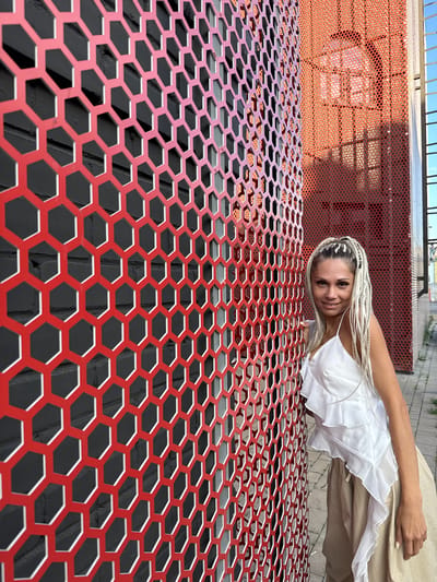 A woman with long, blonde braided hair leans against a red, hexagonal metal screen in an urban environment. She is wearing a white ruffled top and tan pants. The background shows a brick wall behind the screen, and further back, a red building with a similar perforated facade. The ground is paved with gray stones. The scene appears to be outdoors during the daytime, with shadows indicating sunlight. The location is specified as Esbol Rural District, Kazakhstan.
