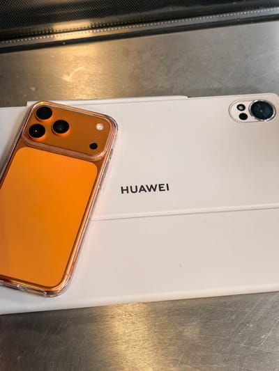 The image shows a close-up, overhead view of a white tablet with the "HUAWEI" logo printed on it, lying on a metallic surface. A transparent-cased orange smartphone is placed on top of the tablet, partially obscuring the logo. The smartphone has a triple-lens camera setup on its rear. The background appears to be a dark, ribbed surface. The scene is indoors and shows no indication of being in Chuchuligovo, Bulgaria.