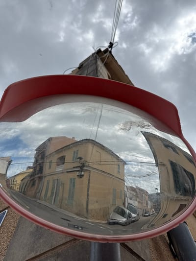 A convex security mirror, framed in red, dominates the foreground in Andratx, Spain. Its scratched and smudged reflective surface displays a distorted view of a narrow, curving asphalt street flanked by multi-story buildings. These structures are predominantly light yellow/beige, with some featuring orange-tiled roofs, closed light blue/green window shutters, and a white balcony railing on a prominent corner building. A silver hatchback car is parked on the street, with additional vehicles visible further down. Overhead power lines are present throughout the reflected scene, and a traditional wall-mounted street lamp is affixed to a corner building. The sky within the reflection is partly cloudy. Above the mirror, against an overcast sky, the upper section of a building or utility structure is visible, from which multiple white electrical or communication cables extend upwards and to the left. No people are present.
