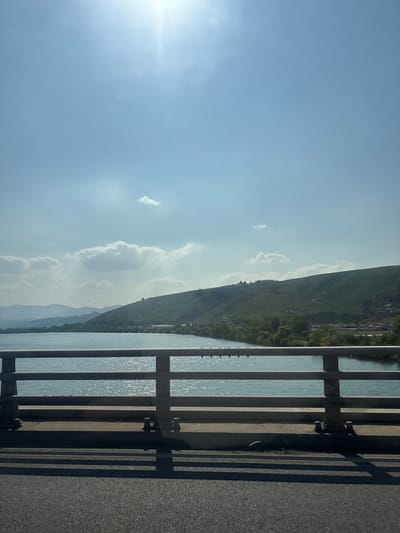 The image captures a view from a bridge overlooking a wide river and a tree-lined shore. The river, reflecting the bright sunlight, has a shimmering, blue surface. Beyond the river, rolling green hills rise, dotted with vegetation and some structures. In the distance, hazy mountains complete the landscape. The sky is a clear blue, with a few scattered white clouds. The foreground of the image shows the asphalt road of the bridge and its railing, casting shadows onto the road surface. The scene appears to be located in the vicinity of Ampuis, France, given the geographical context of the Rhône river and its surrounding hills.