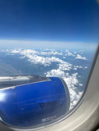 The image is taken from an airplane window, looking out at the landscape below and the sky above. In the foreground, a portion of the airplane's blue engine nacelle is visible. The nacelle is metallic with visible rivets and some warning labels.

The landscape below is a mix of rural and mountainous terrain, with a clear sky dotted with fluffy white clouds. The ground appears to be covered in green vegetation, with some signs of human habitation like roads and scattered buildings visible in the distance.

The sky is a deep blue, transitioning to lighter shades near the horizon. The bright sun is glinting off the engine nacelle, indicating it is daytime and likely sunny.

While the image shows a landscape, it is not possible to definitively determine if it is specifically the city of Viehhausen, Austria, based solely on the visual information provided. The image lacks any identifying landmarks or specific geographical features that would pinpoint its location to that city.