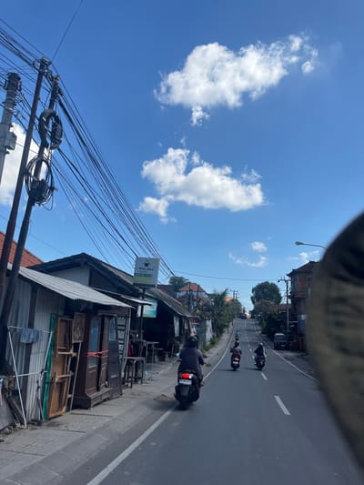 In Denpasar Barat, Indonesia, on a clear day with scattered clouds, a street scene unfolds. The perspective is from a moving vehicle, looking forward along a paved road lined with buildings. On the left, a row of structures with corrugated metal and tiled roofs are visible, featuring wooden furniture and signage. A prominent utility pole with numerous wires crisscrosses the sky above. Several motorcycles are in motion on the road; three are clearly visible in the foreground and midground, with riders wearing helmets. In the distance, more vehicles and buildings are discernible, suggesting a moderately busy urban environment. The overall impression is of a typical street in a tropical city.