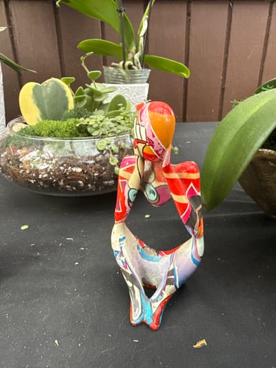 The image shows a still life scene composed of a small, clear glass bowl containing a plant arrangement, alongside a colorful, patterned figurine, set against a backdrop of brown horizontal wooden planks. The plant arrangement features a variety of greenery, including a heart-shaped variegated Hoya leaf, moss, and other trailing plants set in soil. The figurine, predominantly orange, red, and blue, appears to be an abstract sculpture. The surface beneath the arrangement and figurine is a dark, possibly black material. The location of the scene is Davie, United States of America.