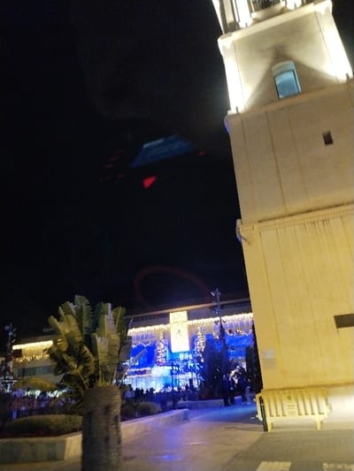 A night shot shows a brightly lit building adorned with blue and white Christmas decorations, including trees and stars. A Ferris wheel in the background glows red. People mill around in front of the building, partially obscured by a palm tree. A tall, light-colored tower stands prominently on the right, its upper portion brightly illuminated. The dark sky contrasts sharply with the festive lights.