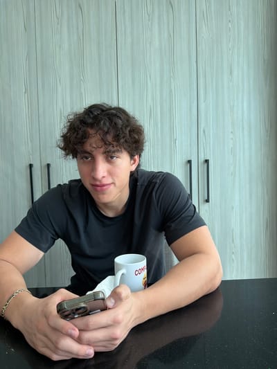 A young man with curly brown hair looks directly at the camera while holding a cell phone in his right hand and a white mug in his left. He is wearing a dark short-sleeved shirt and a bracelet is visible on his right wrist. The background features light wood-grained cabinetry on the left and a dark, reflective countertop on the right. The image is rotated counter-clockwise.
