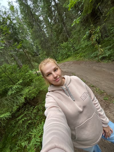 A young woman with blonde hair tied back in a bun is taking a selfie in a forest. She is wearing a light pink hooded sweatshirt and blue jeans. The forest is dense with tall evergreen trees and lush green undergrowth. A dirt path winds through the trees behind her. The woman is smiling slightly at the camera. The location is identified as Anteplevo (Okhotinskoe S/P), Russia.