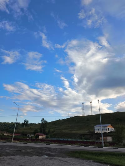The image shows a view of a hilly landscape with a rural settlement in Aya, Russia. In the foreground, there is a paved road with a curb and a gravel shoulder. A lamppost stands on the side of the road. Behind the road, there is a reddish-brown fence. Beyond the fence, several wooden structures with red roofs are visible, some of which appear to be houses or sheds. Further up the hill, a modern house under construction is situated on a slope, with exposed brickwork and unfinished elements. Two tall communication towers are prominent on the crest of the hill. The landscape is covered in green grass and scattered trees. The sky above is a vibrant blue, dotted with fluffy white clouds. The lighting suggests it is either early morning or late afternoon.