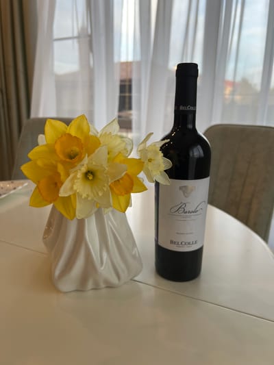 A bottle of Bel Colle Barolo wine lies on a white table, alongside a white vase filled with yellow and white daffodils. The scene is located indoors near a window covered with a sheer white curtain. Parts of a chair are visible.