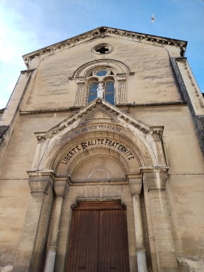 A low-angle shot reveals a stone building under a partly cloudy sky. Above the wooden double doors is an arched stone inscription "LIBERTE EGALITE FRATERNITE" beneath "REPUBLIQUE FRANCAISE". Stone columns frame the entrance. A niche with a statue sits above the entrance, topped by a circular window. The building's facade is textured, and a single bird flies in the sky.