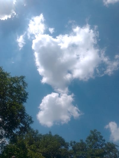 The image shows a bright blue sky with several white, fluffy clouds. The sun is shining brightly, creating a lens flare effect in the upper left corner of the image. In the foreground, there are trees with dark green leaves, some of which are silhouetted against the sky. The trees appear to be healthy and full. There are no people visible in the image. The overall impression is one of a clear, sunny day. This scene is consistent with the natural landscape found in and around Yessentuki, Russia, which is known for its picturesque scenery and clear skies, particularly during the warmer months.