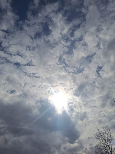 The image captures an outdoor scene in Avdot’ino, Russia, featuring a sky predominantly covered by a mix of stratocumulus and cumulus clouds. The sun is partially visible through a break in the clouds in the lower-middle portion of the frame, emitting a bright, intense light and a prominent lens flare. The cloud cover varies from bright white where the sun penetrates to darker grey in other areas, with patches of clear, dark blue sky visible between the cloud formations, particularly in the upper left. In the lower right quadrant, three parallel utility lines, likely power lines, are observed extending diagonally upwards from the right edge. Below and to the right of these lines, the bare, intricate branches of a deciduous tree are visible, silhouetted against the sky. There are no individuals, structures, or specific actions identifiable within the frame beyond these natural and utility elements.