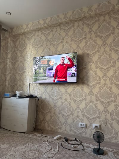 The image depicts an indoor scene, likely a living room or bedroom, in an apartment in the city of Nefteyugansk, Russia. The walls are covered in a beige damask-patterned wallpaper. A large flat-screen television is mounted on the wall, displaying a man in a red jacket pointing upwards, with text in Russian that includes "ЗОЛОТОЙ УРОВЕНЬ" and "Даниил Круговой". Below the TV, on a white cabinet, are some stacked items, including what appears to be a plastic container and a bowl. Cables are visible extending from the TV and a power strip on the floor. To the right, near the wall, are two electrical outlets and a small black portable fan. A light-colored rug with a patterned design covers the floor. The overall impression is of a lived-in space.