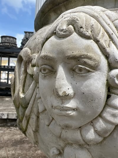 An outdoor, medium-range, eye-level, vertical-format, close-up photograph showcases a weathered, light-gray stone carving of a young woman's face. The carving is part of a larger decorative element, possibly a vase or architectural feature, and is positioned on the left side of the frame, angled slightly towards the viewer. The woman's face is rendered with soft features, including closed eyes, a small nose, and gently parted lips. Her hair is styled in stylized curls and waves that frame her face. The stone exhibits signs of age and exposure to the elements, with visible pitting and discoloration in various areas.
In the background, out of focus, are elements of an outdoor setting. To the left, a large, ornate, dark stone structure with circular decorative patterns is visible, possibly a balustrade or urn. Further behind that, a glimpse of a dark metal fence with rectangular openings and a pale yellow building behind it can be discerned. The sky above is a pale blue with scattered white clouds. The ground appears to be made of stone or concrete. The overall impression is one of an aged architectural detail in an outdoor environment.
The carving's design and the architectural elements in the background suggest a location with classical or historical influences. The mention of Pa Khlok, Thailand, implies the setting is within this specific locale.