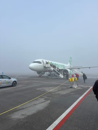 The image depicts an Airbus A320neo airplane, painted in Transavia livery, parked on an airport tarmac in foggy conditions. Passengers are disembarking via mobile stairs connected to the aircraft's front door. A ground service vehicle, labeled "WISAG" is parked nearby. The tarmac has painted red and white lines. The location is at Berlin Brandenburg Airport (BER) in Schönefeld, Germany, based on the plane's registration.