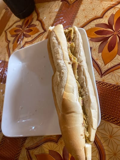 A high-angle, close-up shot presents a substantial sandwich, resembling a sub or baguette, resting on a white, rectangular serving tray. The sandwich is generously filled with a yellow, textured mixture, consistent with scrambled eggs, interspersed with small, reddish-brown pieces that appear to be ham or sausage. The bread itself has a light, slightly golden-brown crust. The white tray shows some minor smudges and possibly crumbs. The sandwich and tray are placed on a decorative surface, likely a tablecloth or mat, adorned with an intricate pattern of geometric shapes and stylized floral motifs in shades of brown, orange, and beige. A dark, indistinct object, possibly part of footwear or a bag, is partially visible in the bottom-left corner. The image captures a ready-to-eat meal or snack, set against a local background, consistent with a setting in Lakka, Sierra Leone.