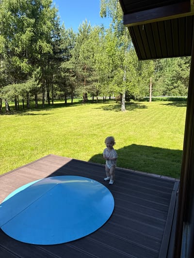 The image depicts an outdoor scene featuring a young child on a wooden deck overlooking an expansive, well-maintained lawn bordered by a variety of trees under a clear blue sky.
The location cannot be forensically confirmed as Копнино, Russia, based solely on the visual content of the image, as no specific landmarks or identifying markers are present. The flora (birch and pine trees) is typical of many forested regions.
A single child, appearing to be a toddler, stands centrally on a dark brown, slatted wooden deck. The child has light, possibly blond or very light brown, curly hair and is wearing a light-colored short-sleeved t-shirt, a white diaper, and white socks with thin blue horizontal stripes. The child faces slightly towards the viewer, exhibiting a subtle smile or happy expression, with hands clasped together at chest level.
On the deck, to the lower left of the child, rests a large, circular, convex object of bright blue color, approximately half the width of the deck. It features a distinct seam or line running across its diameter. Part of the deck, particularly on the right side and under the blue object, is cast in shadow originating from an unseen overhead structure, a portion of which (a dark brown eave or overhang with visible hooks) is visible in the upper right quadrant of the image.
Beyond the deck, a meticulously mowed, bright green lawn stretches into the midground, displaying faint horizontal mowing stripes. The lawn is bordered by a dense line of trees, including deciduous trees with light green foliage, likely birches, and various coniferous trees such as pines. In the far background, additional greenery and a patch of purple flowers are discernible. The sky above is clear and vibrant blue, indicative of fair weather. The distinct shadows suggest bright sunlight, likely during midday or early afternoon.