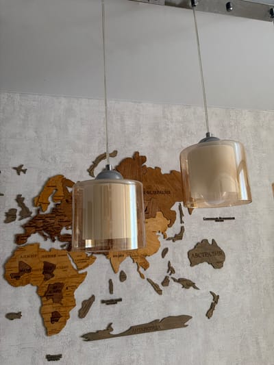 The image displays a wall decorated with a wooden map of the world. Two pendant lights hang from the ceiling, with translucent amber-colored glass shades. The map is made of intricately cut wooden pieces, with country names in Cyrillic text visible on several continents, including Africa, Europe, Asia, and Australia. The lighting is dim, casting a soft glow on the wall. The wall itself has a textured, light gray finish. There are no people visible in the image, nor are there any actions or interactions taking place. The overall scene suggests an interior setting, possibly a room in a home or office. The specific location in Semiluki, Russia, is not evident from the visual information presented in the image.