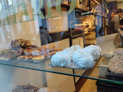 A glass display case is filled with pastries and sweets. Croissants, tarts, and meringues are arranged on metal trays. In the background, reflections on the glass hint at a bustling bakery interior with shelves of bottles and a glimpse of a person behind the counter. The scene is brightly lit, highlighting the textures of the baked goods.