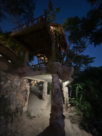 The image depicts an outdoor scene at night, likely a restaurant or lodging with an elevated wooden structure resembling a treehouse or balcony, supported by stone pillars. The architecture incorporates natural elements, with a tree growing through or alongside the structure. A stone wall forms the base of the building on the left, leading to a pathway and stairs. In the background, there are more trees and foliage, suggesting a natural or jungle-like setting. Small lights are visible further up the stairs, hinting at an open area with activity or seating. The sky is dark blue, indicating it is nighttime. The overall atmosphere is rustic and somewhat whimsical, with the interplay of natural and man-made elements. This location is within the city of Karimunjawa, Indonesia.