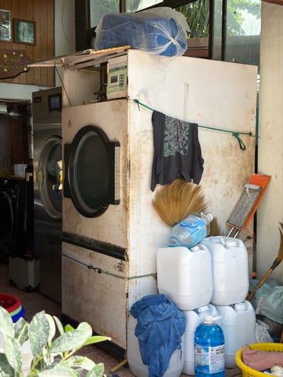 The image depicts an outdoor laundry area on the island of Ko Tao, Thailand, likely in a subdistrict with this name. The scene is dominated by commercial-grade laundry appliances, including two large front-loading washing machines and a dryer. One of the washing machines is stainless steel and has its door open, revealing laundry inside. Next to it is a taller, white dryer unit with its door closed. Above the dryer, a large blue bundle wrapped in plastic is visible.

A dark gray t-shirt with a graphic design is hanging on a green rope strung across the front of the dryer. To the right of the t-shirt, a broom with a natural bristle head rests against the wall. Below the broom, an orange dustpan is propped up, filled with what appears to be dust and debris.

A collection of white plastic containers, some stacked, are situated at the base of the dryer. One of these containers has a blue bottle of liquid detergent placed on top. A blue towel is folded and placed on top of one of the stacked containers. To the far right, a yellow laundry basket contains some pink fabric. In the foreground, a potted plant with lush green leaves is visible. The overall impression is of a functional but somewhat cluttered laundry service area.