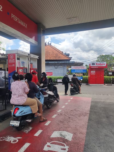 The image captures a bustling scene at a Pertamina gas station in Bedulu, Indonesia. Under the red and white "PERTAMAX" sign, people on motorbikes gather for fuel. A woman in a light pink, bird-patterned shirt sits on a light blue scooter with two passengers, receiving service from a gas station attendant in a red shirt. Two other people on motorbikes are waiting in line.

The foreground showcases the designated lane for motorbikes, marked with red paint and white car silhouettes. In the background is a red "Enduro Motor Service" station and a sign warning against smoking. A building with a red tile roof is visible beyond the station and signage. Powerlines run across the sky, which is filled with clouds.
