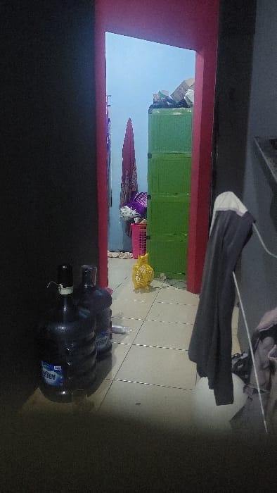 The image shows an open doorway in a building in Ciampea, Indonesia (latitude: -6.56, longitude: 106.716). The doorway leads into another room, which is partially visible. The room appears to be a storage area or utility room, with several objects visible. There is a green plastic storage cabinet on the right side of the room, filled with various items. A pink plastic bin is on the left side of the room, containing items. A yellow plastic object resembling a toy or storage container is in the center of the room.  A long, dark-colored garment is hanging on a hook on the left wall of the doorway. Two large, plastic water jugs are stacked on the floor just outside the doorway, with the label "Aqua" visible. The floor is tiled and appears to be clean. No people are visible in the image. The lighting is dim, suggesting either dusk or the presence of only artificial lights.  The doorway is framed by a red wall on the left side and a grey wall on the right side.  The room beyond the doorway is brightly lit, suggesting the presence of a window or other light source.  The overall impression is of a modest, functional space.
