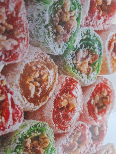 The image shows a close-up, slightly blurred view of a pile of colorful Turkish delight candies. The candies are round and appear to be rolled or twisted, with a filling of nuts, possibly walnuts or pistachios. They are coated in a white powdery substance, likely powdered sugar or shredded coconut. The dominant colors of the candies are red, green, and a light orange or brown. The candies are arranged haphazardly, with some stacked on top of others, filling the frame. The lighting is bright and diffused, suggesting an indoor setting. There are no people, text, or clear environmental cues to indicate a specific time of day, weather, or location beyond the provided context of Dolna Malina, Bulgaria. The focus is entirely on the texture and color of the sweets.