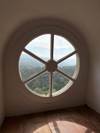 A high-angle, full shot captures a circular window in a room with brick flooring. The window, divided into six sections by white bars meeting at a central hub, looks out onto a forested landscape with hills and a sky dotted with clouds. The walls of the room are off-white, and shadows fall across the brick floor. This scene likely takes place at Quinta da Regaleira, a historic estate located in Sintra, Portugal.