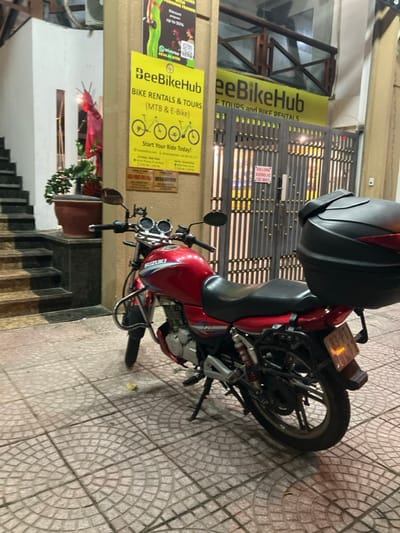 An outdoor, nighttime scene in Ngũ Hành Sơn Ward, Vietnam, depicts a red Suzuki motorcycle parked on a tiled sidewalk in front of a building entrance. The motorcycle, equipped with a black seat, chrome crash bars, and a black hard-shell top case, faces left with its kickstand deployed. Its rear license plate, partially visible, begins with "43-P1". The sidewalk features square gray tiles with circular relief patterns. To the left of the motorcycle, a dark-colored staircase ascends next to a large brown potted plant containing green foliage and red decorative items. To the right of the motorcycle, a light-colored building pillar displays two vertical yellow banners. The lower banner advertises "BeeBikeHub BIKE RENTALS & TOURS (MTB & E-BIKE)" with bicycle graphics. The upper banner is a "PERSONAL TRAINER" advertisement with a WhatsApp number and QR code. Adjacent to the pillar is a dark gray metal gate, partially revealing an illuminated interior. A white sign with red text on the gate reads "VUI LÒNG XUỐNG XE TẮT MÁY" (Please get off the bike and turn off the engine). Above the gate, another yellow "BeeBikeHub" sign is visible. No individuals are present in the scene.