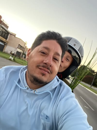 A selfie-style photograph captures two individuals in City Bell, Argentina, during late afternoon or early evening. In the foreground, a man with dark hair, a mustache, and a goatee, wearing a light blue hooded sweatshirt with "The North Face" logo, looks towards the camera with a slight smile. Behind him and to his right, a second person, mostly obscured, wears a black motorcycle helmet featuring a silver stripe and the number "721". This individual is leaning in towards the man's right cheek with pursed lips. A bundle of long, green plant stalks extends upwards from behind the helmeted person. The background shows a paved road with a white lane marking on the right, flanked by green vegetation. On the left, several buildings are visible, including one displaying a "DUNLOP SERVICIOS" sign. The sky is bright and clear, illuminated by a warm, low-angle light. The scene implies motion, consistent with being on a moving vehicle like a motorcycle.