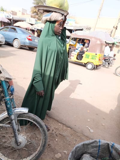A woman in a green hijab and robe balances produce on her head. She stands on a street lined with cars, motorcycles, and a three-wheeled vehicle with the number "TAN 3277". A bicycle wheel is in the foreground to the left, and a woven bag is visible in the lower right corner. Other people and businesses with awnings are visible in the background. The street is dusty and appears to be located in an area of Africa.