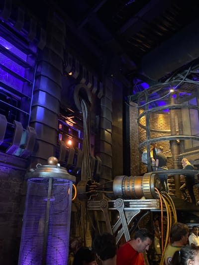 The image depicts an interior space with industrial theming, possibly a ride or attraction. A large metal cage structure, likely a component of the ride, is visible with people inside. Other metal structures and pipes are present, illuminated by blue and orange lighting. Some people are visible standing nearby, observing the scene. Based on the 'Rust' query, this is likely at the Europa-Park theme park in Rust, Germany.