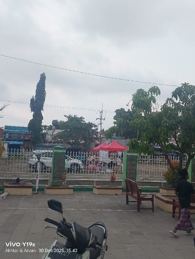 The image depicts an outdoor public area in Jagapura Kidul, Indonesia, under an overcast sky at approximately 15:42 on December 30, 2025. The foreground is a paved surface composed of light gray square tiles. A white and black scooter is parked on the left side of this paved area, facing slightly left. To the right, dark brown wooden benches are visible, along with planter boxes featuring green and yellow trim, containing small plants.
A white metal fence with decorative elements and pillars accented with green and dark gray separates the paved area from a street or market. Beyond this fence, in the midground, multiple red canopy tents are erected, indicating a market or temporary gathering. Several vehicles, including at least one white pickup truck, are parked or moving near these tents. Indistinct human figures are visible within and around the tent area, suggesting activity. Buildings with various colored facades, including blue and light brown, line the background street. Utility poles and overhead power lines are visible against the sky.
On the far right of the paved area, a single individual is walking towards the right. This person is attired in a dark-colored top and a red and blue plaid sarong-like lower garment. Trees are present throughout the scene; a tall, slender tree stands on the left background, while a tree with dense green foliage is prominent on the mid-right. The sky is uniformly light gray, indicating cloudy weather conditions.