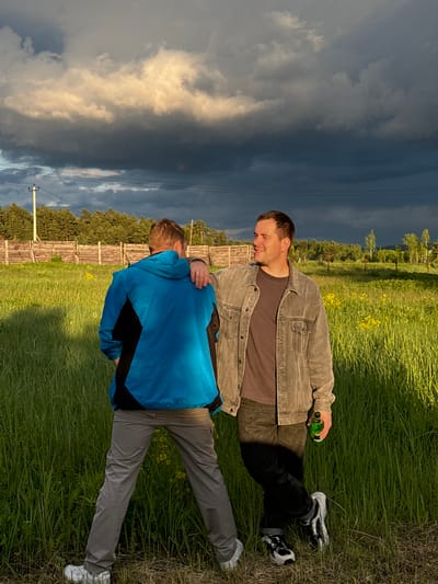 Two men are lying on a grassy field in Vysokinichi, Russia. One man wears a grey jacket, brown shirt, and black pants, holding a green bottle. The other man wears a blue and black jacket, and grey pants. The man in the grey jacket has his hand on the other man's shoulder. A wooden fence runs along the edge of the field. A dark, cloudy sky fills the background. Power lines and a utility pole are visible.