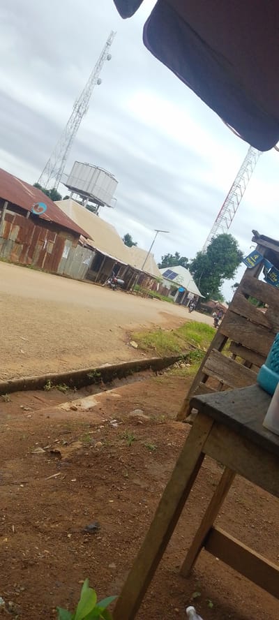 This media file depicts an outdoor scene in Baissa, Nigeria, under an overcast sky, suggesting either early morning, late afternoon, or a cloudy day. The foreground is dominated by a wooden structure, possibly a market stall or makeshift seating area, constructed from rough-hewn planks. To the right of this structure, a blue plastic container or bottle is visible.

The midground reveals a dirt road lined with a shallow, overgrown drainage ditch. Along the road, a row of simple, tin-roofed structures, likely shops or homes, are visible. Their corrugated metal walls appear aged and rusted in places. A water tower stands prominently behind some of these buildings, supported by a concrete structure. Two tall communication towers, one with red and white sections, are visible in the background, stretching towards the sky.

Several motorcycles are parked or being ridden on the road, indicating a place of some local activity. One motorcycle is parked in front of a shop, while others are seen further down the road. A few people are present, appearing as small figures in the distance, suggesting a casual, everyday atmosphere rather than a special event. The overall impression is of a rural or semi-urban setting in Nigeria, with a focus on functional, practical infrastructure and buildings. The cloudy weather contributes to a subdued but not necessarily somber mood. No distinct text is clearly legible in the image, although some signs might be present on the structures that are too small or blurry to read.