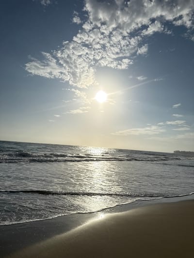 The image depicts a beach scene in Cabarete, Dominican Republic. The foreground shows wet sand, transitioning into shallow ocean waves reflecting sunlight. The background features a clear, blue sky with scattered white clouds. The sun shines brightly, creating a hazy glare and illuminating the entire scene. A treeline is visible on the horizon.