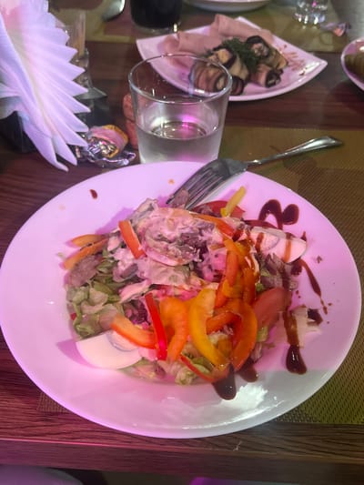 The image shows a table setting at a restaurant. In the foreground, a white plate holds a salad with lettuce, tomatoes, bell peppers, and what appears to be hard-boiled eggs and meat. The salad is drizzled with a dark sauce. To the left of the plate, a fork rests on the table, and a glass is partially visible. In the background, a decorative dish of rolled eggplant appetizers is also on the table. The setting appears to be indoors, with a wooden table and a woven placemat visible. The lighting in the image is somewhat dim, with a pinkish hue cast over the food. The overall impression is of a meal being served in a restaurant. There is no visible text.