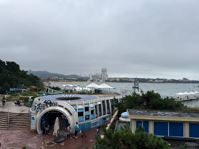 This image captures a coastal scene on an overcast day, specifically depicting the Qingdao Seal Museum in Shinan District, Qingdao, China.
In the foreground, the "SEAL MUSEUM" (海豹馆) is a dominant structure, semi-circular in design with a light blue and white exterior. Its roof is extensively covered with radially arranged solar panels. A large arched entrance is visible on the left side of the building, where approximately six individuals are observed entering or exiting. One person is pushing a stroller. A dark paved plaza extends in front of the museum, and to its left, a multi-tiered stone stairway leads up to a higher vantage point where a few people are seated or standing. To the lower right, part of a rectangular building with a dark roof and blue-paneled exterior is visible, partially obscured by green coniferous trees.
The midground features a large body of water, likely Fushan Bay, with a lengthy pier or floating walkway extending from the right, supporting multiple white, tent-covered structures. A prominent metal framework, possibly a diving platform or suspended structure, is positioned over the water. Along the distant shoreline, a sandy beach is visible, populated with numerous white beach umbrellas and tents, indicating a recreational area.
The background reveals a dense urban landscape characterized by numerous buildings, including a cluster of high-rise structures, one tall white skyscraper with a distinctive finial. Beyond the city, rolling green hills and mountains are visible under a uniformly gray, overcast sky, indicating cloudy weather conditions.