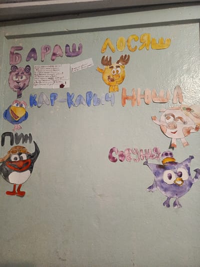 The image displays a colorful, hand-drawn collage of cartoon characters and text, likely a decoration for a children's event or classroom. The artwork is affixed to a light blue, textured wall, possibly a door or bulletin board. The characters include a purple bear, a yellow moose, a blue bird, a penguin wearing a pilot's hat, a pink pig with a hat, and a purple owl-like creature. Large, painted letters are positioned above and around the characters, spelling out what appear to be names or titles. For instance, the word "БАРАШ" is prominently displayed in purple letters, and "ЛОСЯШ" in yellow. Below these, "КАР-КАРЫЧ ННШ" is written in blue and orange/pink letters. The penguin is labeled "ПИН" in black letters. The pig is labeled "СОВУНЬЯ" in purple letters. There are also handwritten notes in Russian on pieces of paper attached to the wall. One note features a drawing of a flower and what appears to be a poem or message in Russian, with a red ink smudge. The overall aesthetic is childlike and whimsical. This image was taken in Meget, Russia, a city in the Irkutsk Oblast.