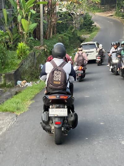 The image captures a line of motorbikes on a curved road in Singakerta, Indonesia. The focus is on the rear of the closest motorbike, displaying its license plate "DK3971KBX" with an expiration date of April 2030, and Yamaha branding. The rider wears a helmet, a white shirt, and a backpack with the logo "DENIM." The road has a grassy verge on the right side with visible litter. In the distance, other motorbikes and a white van follow the curve of the road. The surrounding environment is lush with vegetation, including banana trees.