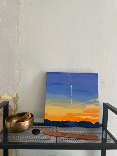 A small, square painting of a sunset hangs on a textured, off-white wall, slightly left of center. The painting depicts a gradient of colors, starting with a deep blue sky at the top, transitioning to shades of orange and yellow at the horizon, with a dark blue silhouette of mountains at the very bottom. A thin, vertical white streak, possibly a contrail, is visible in the upper part of the sky. To the left of the painting, on a black metal shelf, sits a brass singing bowl containing a wooden mallet. Next to the bowl, there is a dark, smooth stone and a wooden incense holder with partially burned incense sticks. The shelf is composed of a mesh grid. The wall behind the shelf shows signs of wear and tear, with peeling paint and visible cracks, particularly on the left side. The overall scene suggests an indoor setting, possibly a room with an artistic or meditative theme. The image does not contain any people or depict any specific urban location within Kopeysk, Russia.