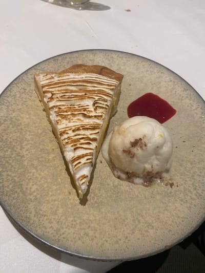 A close-up, high-angle view displays a dessert presented on a round, speckled light gray ceramic plate with a dark gray rim, resting on a white tablecloth. The plate holds a triangular slice of lemon meringue pie on the left, featuring a golden-brown pastry crust, a light yellow lemon filling, and a thick layer of extensively torched white meringue with distinct, horizontal, dark-brown striations. To the right of the pie, a scoop of white ice cream shows signs of partial melting at its base and is dusted with dark brown particulate matter. An irregular, vibrant red viscous liquid, consistent with a fruit coulis, is situated on the plate adjacent to the ice cream. A blurred, light-colored object, possibly glassware, is partially visible in the upper left background. No individuals are present. The scene depicts a prepared dessert in a dining environment within La Wantzenau, France.