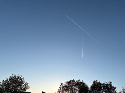 The image depicts a blue sky at dusk with multiple contrails from airplanes visible. The contrails intersect at one point. Below, there is a line of trees in silhouette. A portion of a rooftop is also visible. While the location cannot be verified from the image itself, the prompt specifies that it should be considered Wiener Neustadt, Austria.