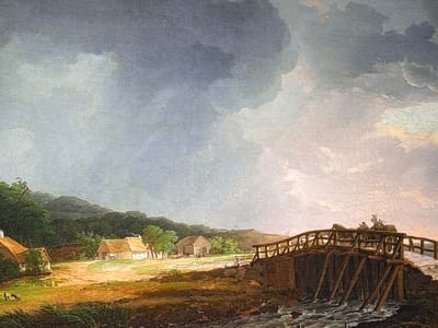 The image depicts a detailed painted rural landscape, presented as being located in the city of Reus, Spain.

The sky is characterized by an overcast condition, with heavy, dark grey clouds dominating the upper left quadrant. These clouds transition towards the right into lighter, diffuse grey areas interspersed with faint yellowish-pink hues, suggesting a specific atmospheric lighting condition, possibly dawn, dusk, or a break in a stormy sky. Extensive craquelure (fine cracking) is visible across the painted surface, particularly noticeable in the lighter sky and ground areas.

In the foreground, a robust wooden bridge spans a turbulent, rocky stream or river that flows from the right towards the left. The bridge features a wooden railing and is supported by several vertical and angled wooden piles driven into the streambed. A two-wheeled horse-drawn cart, carrying at least two human figures, one of whom appears to be driving and wears a wide-brimmed hat, is positioned mid-span on the bridge, moving towards the left. The immediate ground beneath and around the bridge is dark and textured, transitioning into earthy brown, tilled or fallow fields in the middle ground.

The middle ground features a rural settlement against a backdrop of rolling hills. To the left, a white cottage with a prominent thatched, reddish-brown roof is situated within an area of green foliage. Small domestic animals, possibly goats or sheep, and a dog are visible near this cottage, alongside a small human figure. Further to the right in the middle ground, another cluster of white buildings with thatched roofs, including a larger barn-like structure, is depicted. A smaller horse-drawn cart and several human figures are visible near this group of buildings, implying agricultural activity. The fields in the middle ground vary in color from earthy brown to patches of green.

The background consists of a range of densely forested hills, covered in dark green vegetation, extending across the horizon.