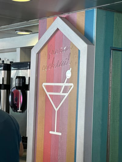 The scene depicts an interior view within a commercial establishment in Puerto del Rosario, Spain. Central to the frame is a white-framed, house-shaped sign. The sign's background consists of vertical planks in pastel colors including pink, orange, light brown, purple, and light blue. Emblazoned on this background are the metallic, cursive words "frozen cocktail," positioned above a white outline graphic of a martini glass with a circular garnish on a stick.

To the right of the central sign, a wall continues with vertical planks in shades of green and teal. To the left, a beverage service counter features at least two commercial stainless steel dispensers. One dispenser has a black top and a clear section displaying a red liquid, with a white dispensing spigot. Multiple bottles are visible on a shelf situated above the dispensers.

In the lower-left quadrant, the upper back of an individual, wearing a dark-colored shirt, is partially visible, facing away from the camera. The ceiling, visible at the top, is light-colored and equipped with recessed circular lights. No active interactions between individuals or objects are depicted.