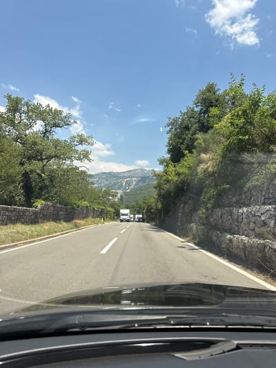 The image depicts a scenic drive through a mountainous region in Podličak, Montenegro. A paved road with white lane markings curves through the landscape. On the left side of the road, a steep, rocky embankment is covered with lush green trees and vegetation. The sky is a clear, vibrant blue with a few scattered white clouds. In the distance, a mountain range is visible, shrouded in a slight haze.
Several vehicles are traveling on the road, including a white van or camper and a darker colored vehicle. The perspective suggests the image was taken from inside another vehicle, likely the passenger seat or rear, looking out towards the road and the surrounding scenery. A portion of the vehicle's interior, including the side mirror and door frame, is visible on the right side of the frame.
The time of day appears to be daytime, likely late morning or early afternoon, given the brightness and the quality of the light. The weather seems pleasant, with clear skies indicating fair conditions for travel. The overall impression is one of a beautiful and serene journey through a natural landscape. There is no visible text in the image.