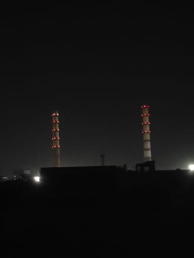 The image shows a night view of the Ust-Kamenogorsk Thermal Power Plant in Oskemen, Kazakhstan. Two tall, industrial chimneys with red lights illuminating their upper sections are prominently visible against the dark sky. The chimneys are cylindrical and appear to be made of concrete or brick. Below the chimneys, a dark silhouette of the power plant's structure is visible, along with some dimly lit lights from other parts of the facility and what appears to be a large, dark building or wall in the foreground. The overall environment is industrial and appears to be a clear night. No people or specific actions are depicted.