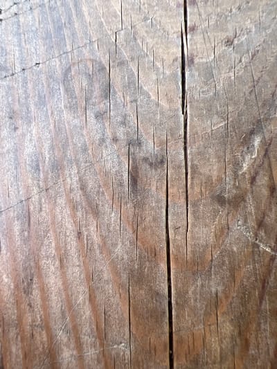 The image is a close-up, top-down view of a weathered wooden surface. The wood grain is prominent, running vertically and diagonally across the frame. Numerous fine cracks and fissures are visible, indicating age and wear. The color of the wood ranges from light brown to a darker, aged brown with some grayish tones. There is a distinct, deep crack running vertically near the center of the image, widening slightly towards the top. The texture appears rough and splintered. The lighting suggests an indoor or shaded outdoor environment, casting subtle shadows that accentuate the wood's texture and imperfections. No people, animals, or other recognizable objects are present. The image provides no information about the city of Ben-Gurion Airport, Israel.