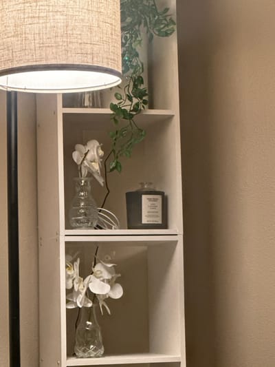 A white, three-compartment shelf holds decorative items. The left compartment contains greenery. The middle compartment features a dark bottle with a label, a glass bottle with white flowers, and electrical wiring. The right compartment has a similar glass bottle with white orchids. A lampshade partially obscures the left compartment. A black rod is positioned below the shelf. The background is a solid, light beige color.